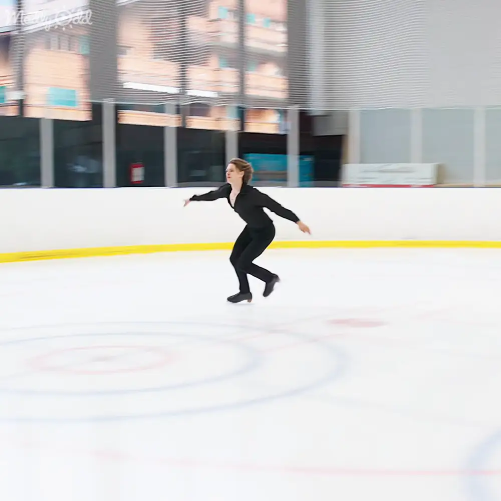 A young adult female figure skater in a black leotard and figure skates on an indoor ice rink with natural light coming through large windows.