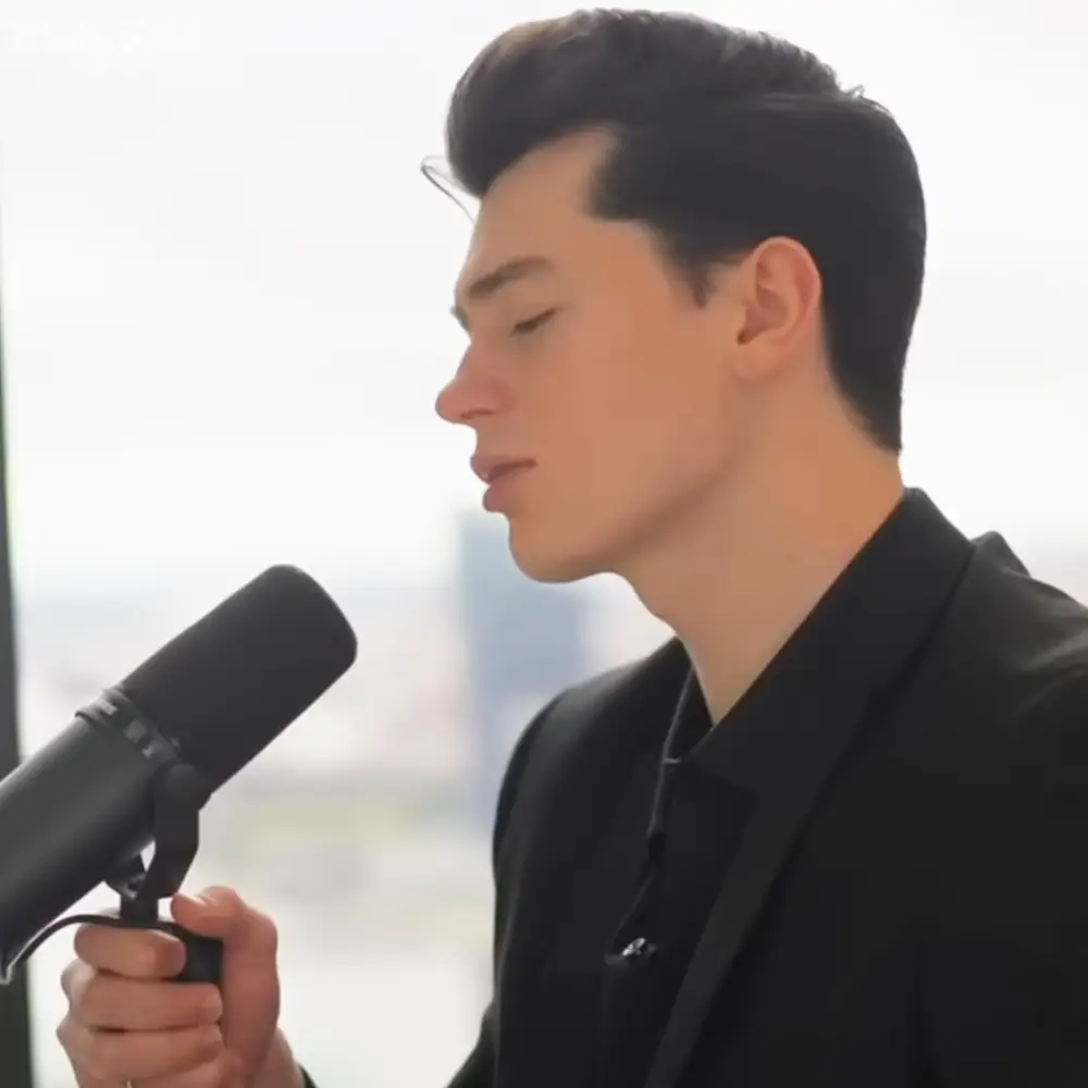 A young adult male in a fitted black blazer with lapels, collared shirt under, holding a microphone with pop filter indoors during daytime.