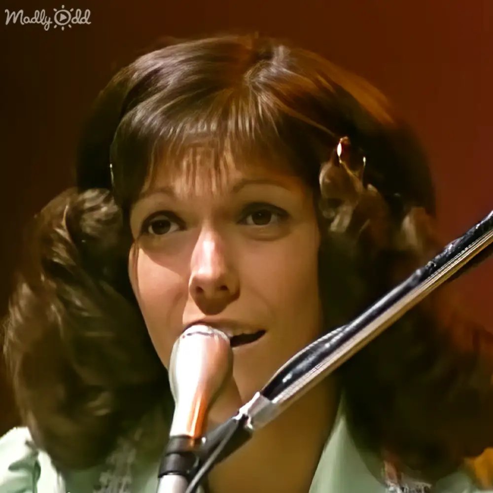 A young adult female wearing a light green, relaxed top performs into a microphone at an indoor stage setting with focused lighting.