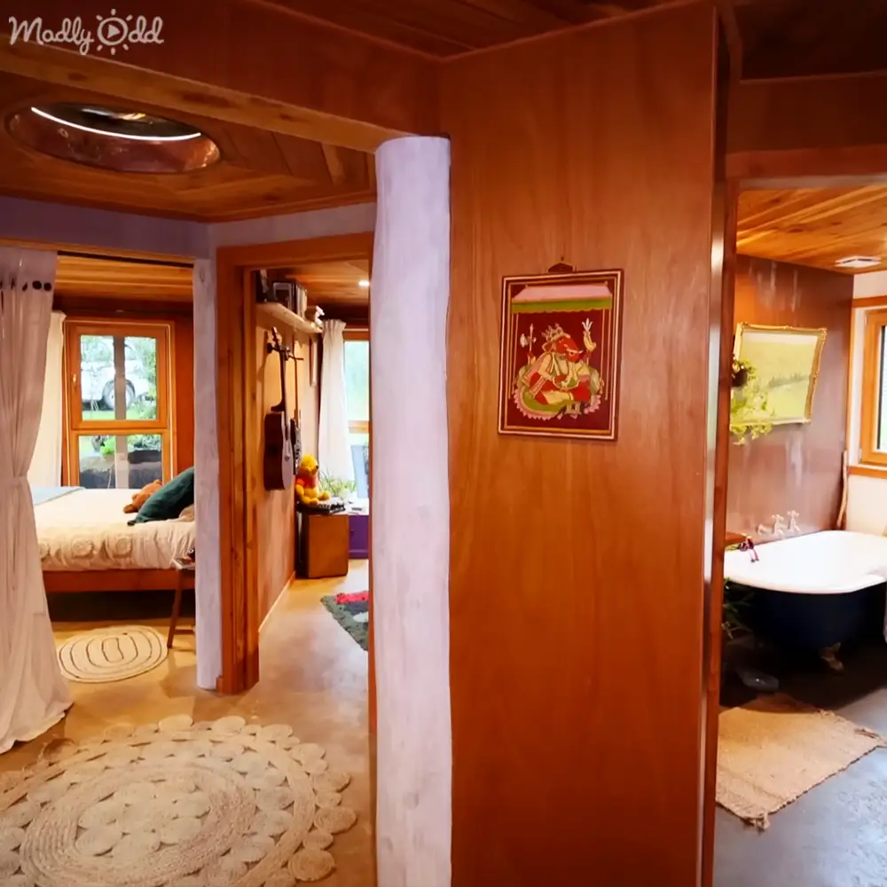 Cozy indoor space with rustic modern decor; a bed with white bedding on left, dark blue bathtub on right, framed pictures of deities and landscapes hanging on walls.