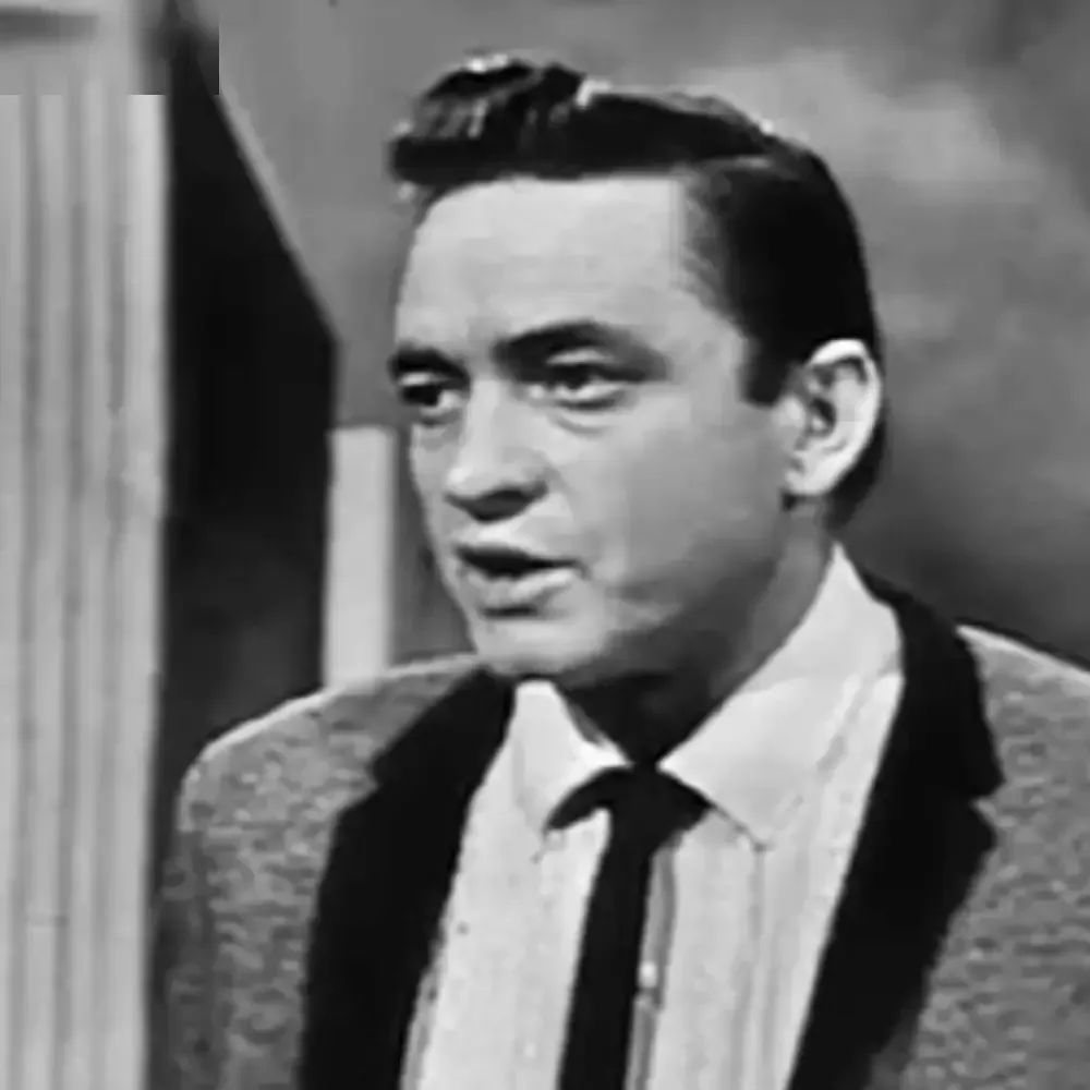 An adult male in a patterned suit jacket, white shirt, and dark tie stands indoors with curtains visible on one side.