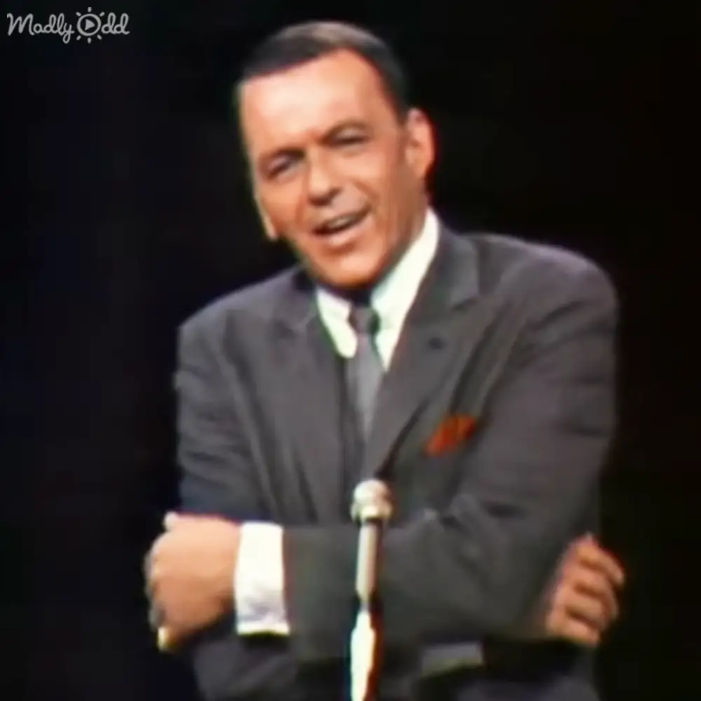 A middle-aged man in a dark gray suit with an orange pocket square, standing indoors on stage.