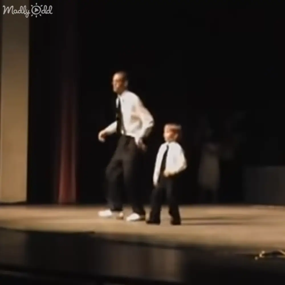 An adult male singer and a child in formal attire perform on an indoor stage during what appears to be a school play or community theater production with dark-toned curtains as the backdrop.