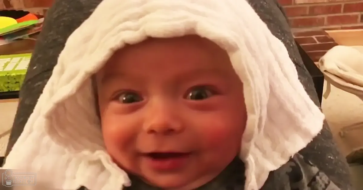 An infant wearing a white hooded garment indoors with a brick wall and folded clothes nearby.