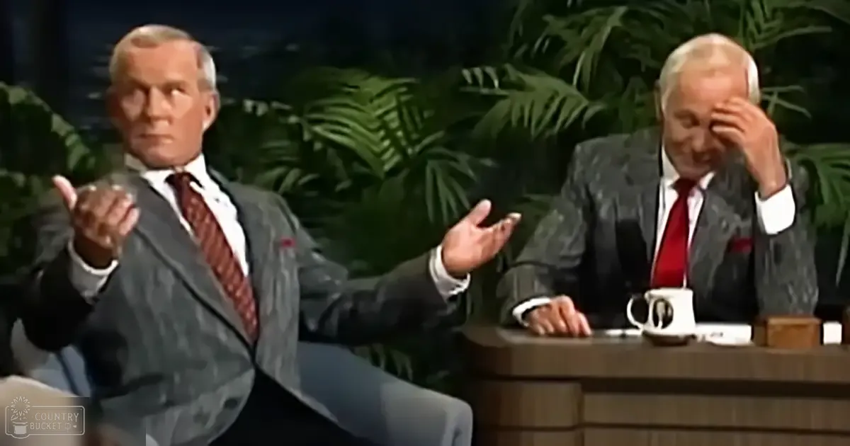 Two middle-aged men seated at a desk in an indoor talk show set with lush greenery in the background.