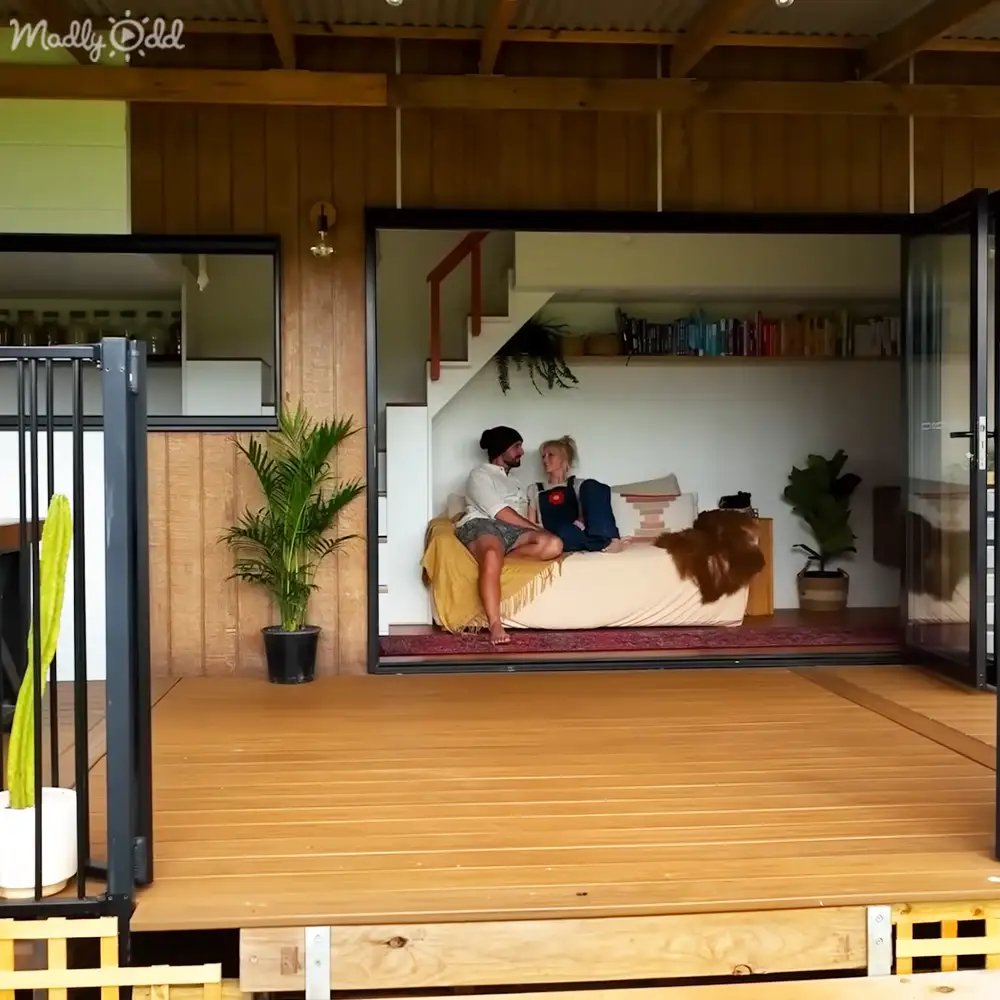 An adult male and female couple sitting on a white couch in a modern, open-concept living space with large glass doors, surrounded by natural light and earthy tones.