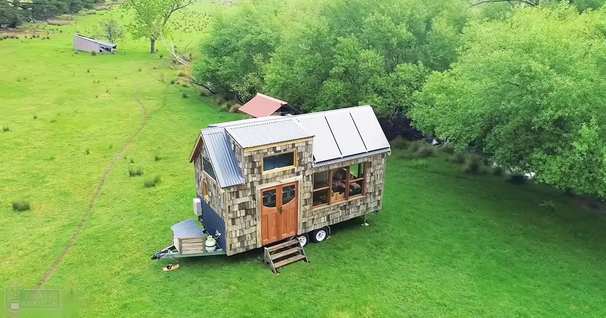 A rural area with a tiny house with a shingled exterior, metal roof, and solar panels surrounded by lush greenery in spring or summer daylight.