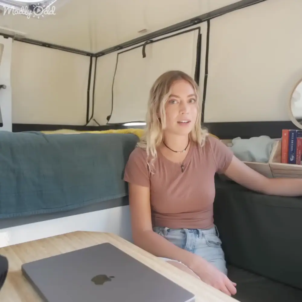 A young adult female sits inside a camper van, wearing a muted pink v-neck t-shirt with blue bedding and yellow pillow behind her.