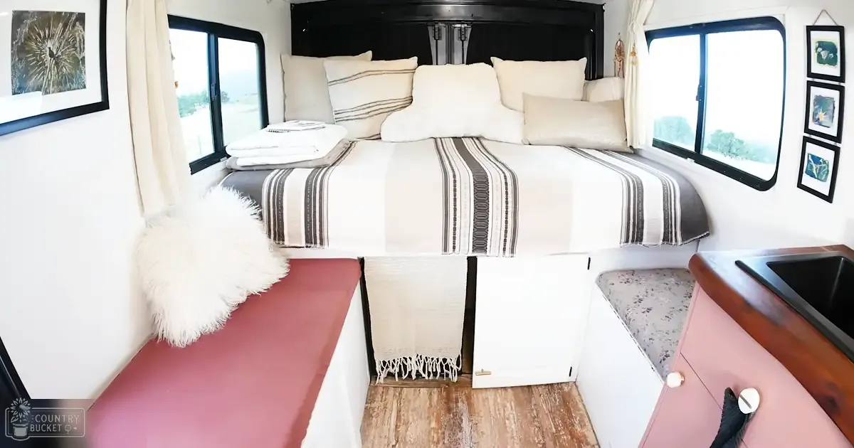 A cozy RV or camper van interior with a dog near the bottom right, framed artwork on walls, pink cushions, white pillows, and natural light from large windows suggesting daytime in spring or summer.