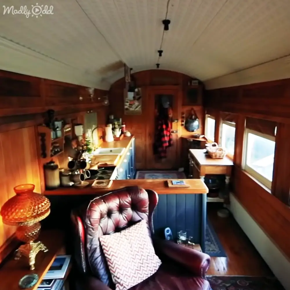 A cozy, vintage-style interior with a red leather armchair, wooden table with a red glass lampshade, blue cabinets in a small kitchenette area, and warm-toned wooden paneling. Daylight streams through the windows in this lived-in space.