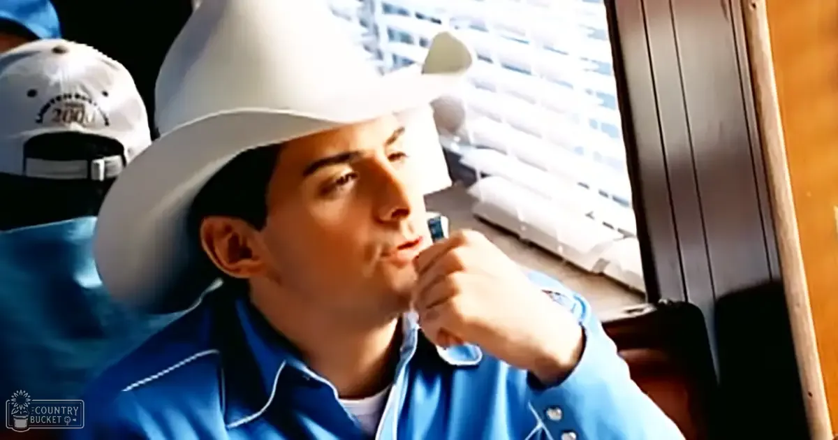 A young adult male wearing a blue button-up shirt with white stitching indoors, seated by a window with blinds partially open.