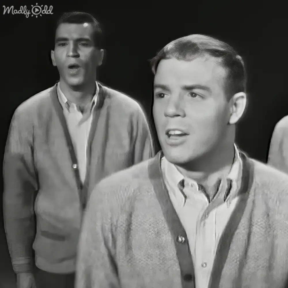 Two young adult males in a dimly lit indoor setting, wearing light gray cardigan sweaters over button-down shirts with subtle patterns under relaxed and casual cuts.