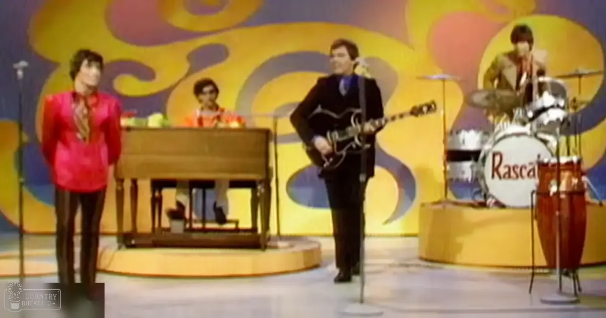 A vibrant, psychedelic-style backdrop with swirling colors frames a stage performance with musicians from the late 1960s or early 1970s, including an adult male singer in a bright red vest and dark pants, another playing a keyboard, a guitarist in a dark suit jacket, and a drummer in a light-colored suit. The setting is an indoor performance space with microphones and a grand piano on stage.