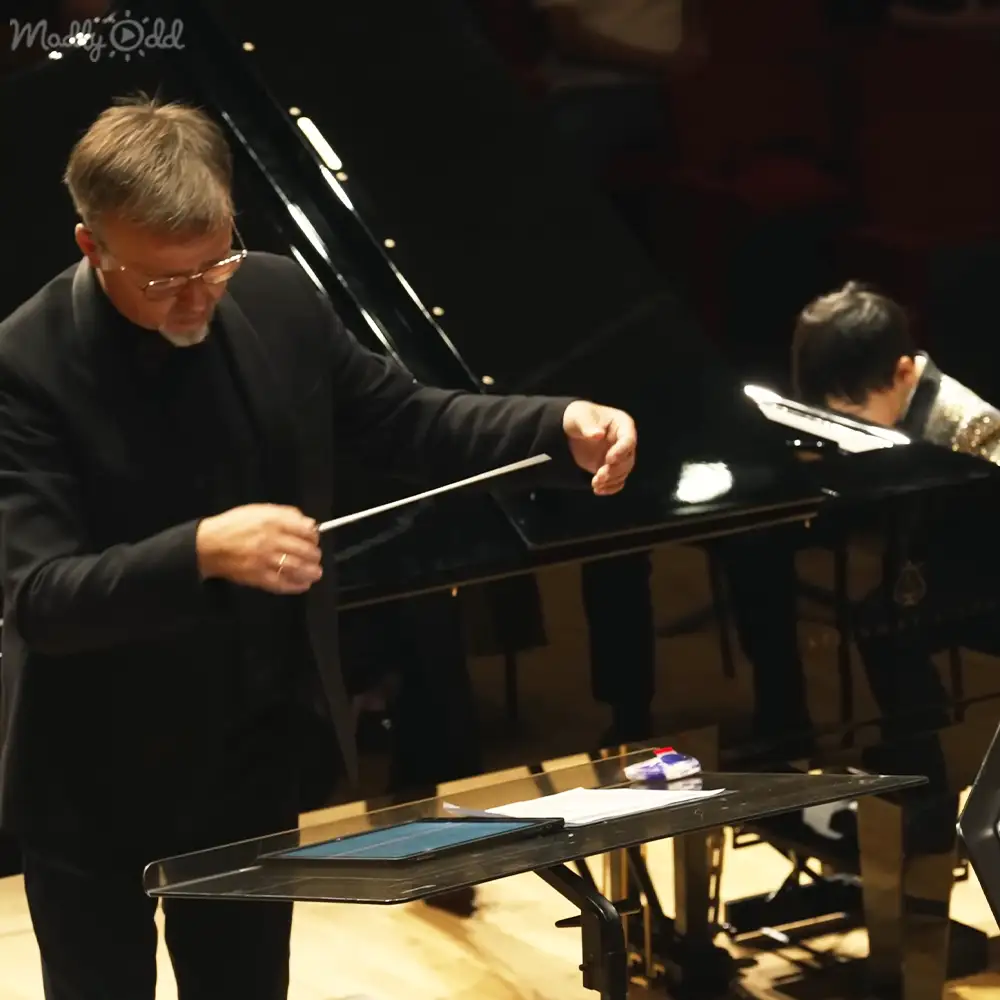 Middle-aged male conductor in a black suit conducting an orchestra in an indoor concert hall with wooden flooring and stage lighting.