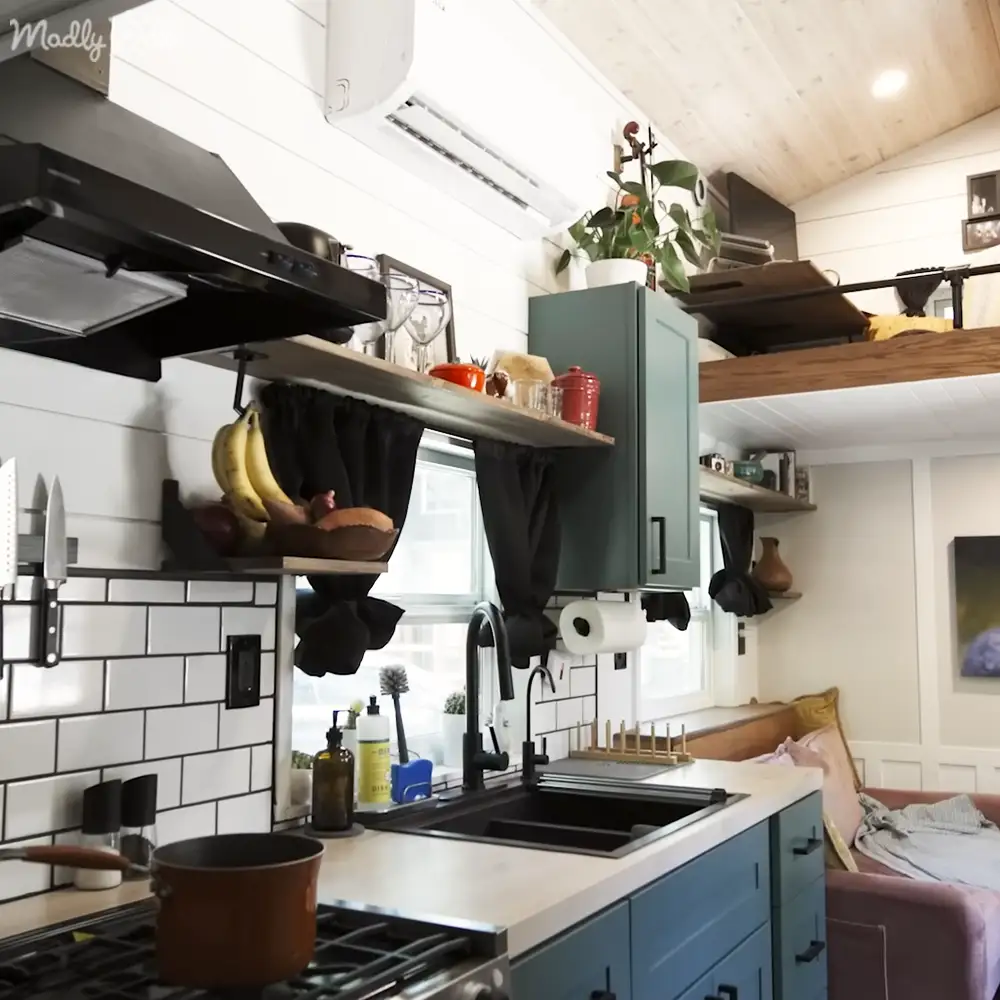 A small home or cabin interior with a gas stove, sink area, blue cabinets, white countertop, and natural light from a window. A couch with a blanket is visible in the background.