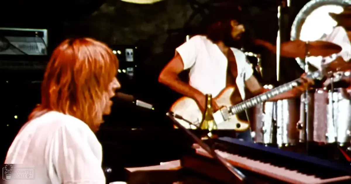 Adult male singer with long reddish-brown hair in a white shirt, accompanied by another adult male with dark skin and shoulder-length hair in a loose-fitting white t-shirt on stage during what appears to be an evening performance.