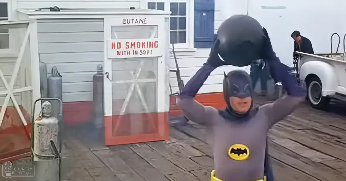 A toddler male child dressed as Batman at an old-fashioned gas station with a 'Butane' sign and wooden dock-like background.