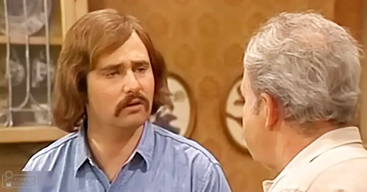 Middle-aged man with long hair and mustache wearing a light blue button-up shirt indoors in what looks like a home setting, accompanied by an older adult male in a white collared shirt. Background includes decorative wallpaper and a shelf holding indistinct items.