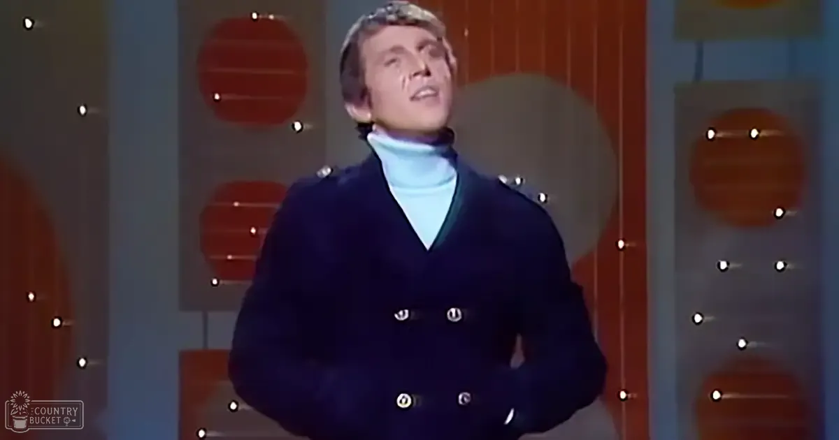 A young adult male wearing a dark blue double-breasted coat and light blue turtleneck sweater stands indoors against circular illuminated elements suggesting a stage or studio setting.