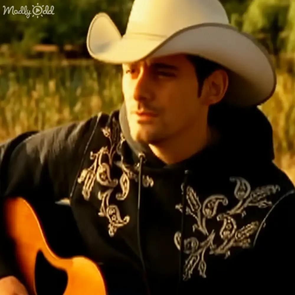 A young adult male wearing a black shirt with intricate white embroidery near water, playing guitar and wearing a white cowboy hat.