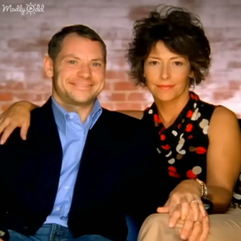 A middle-aged couple in an indoor setting with a brick wall backdrop, wearing casual and summer-appropriate clothing.