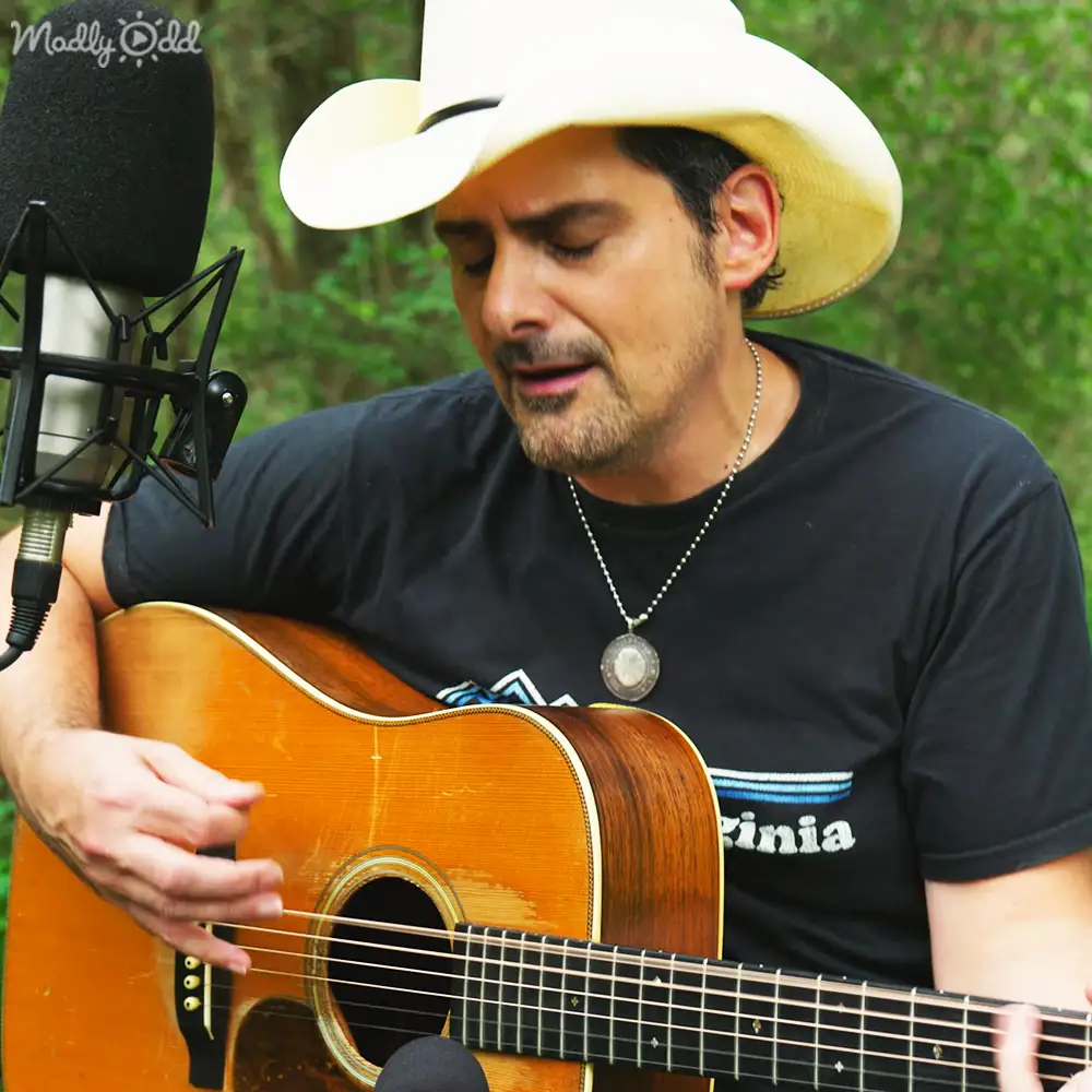 A middle-aged man playing an acoustic guitar outdoors, wearing a black T-shirt with 'Virginia' and a white cowboy hat. Microphone nearby suggests recording.