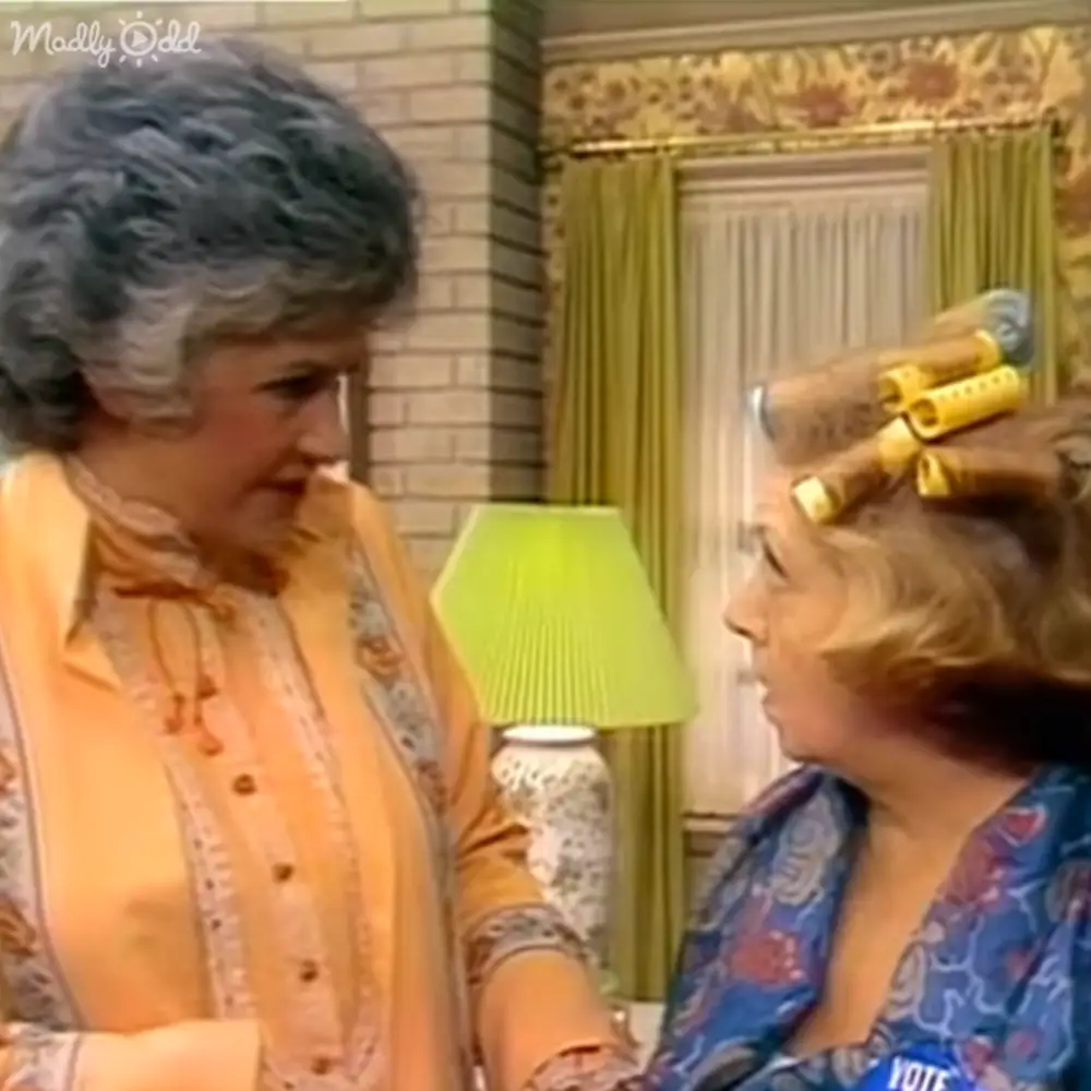 Two older adult females in a home setting, wearing patterned blouses and dresses with floral designs. Curtains and a textured wall suggest a cozy interior.