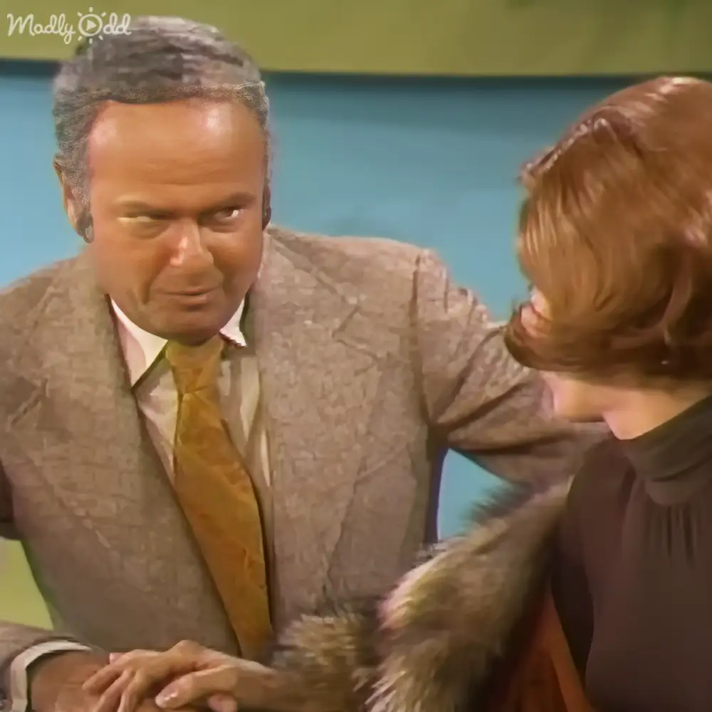 An adult male in a light brown suit jacket with a subtle pattern, mustard-yellow tie, and dark-colored turtleneck sweater stands indoors at what appears to be an event or talk show. A small juvenile terrier mix dog with brown and white fur is present nearby.