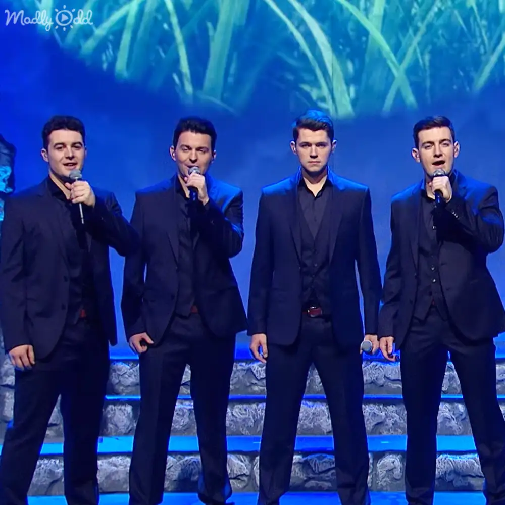A singing quartet in formal dark suits performing on an indoor stage with blue lighting and a green foliage backdrop.