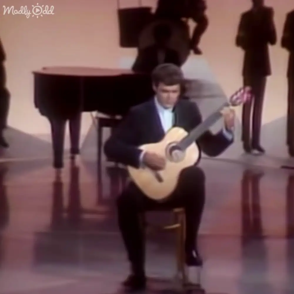 A young adult male plays an acoustic guitar in a formal indoor setting with silhouettes of other individuals and a grand piano, under artificial lighting.