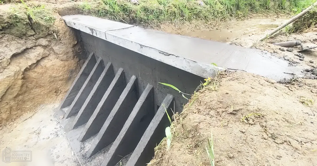 An outdoor rural setting with a concrete structure and steps leading to what appears to be a drainage channel surrounded by green vegetation under natural daylight in spring or summer.
