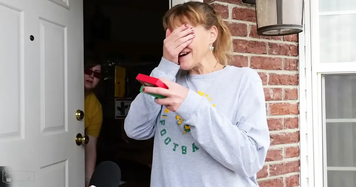 A middle-aged woman wearing a gray sweatshirt with 'FOOTBA' text and yellow graphic stands near a white door, holding a red and green object in her hand. A young adult to middle-aged female in a yellow top stands nearby.