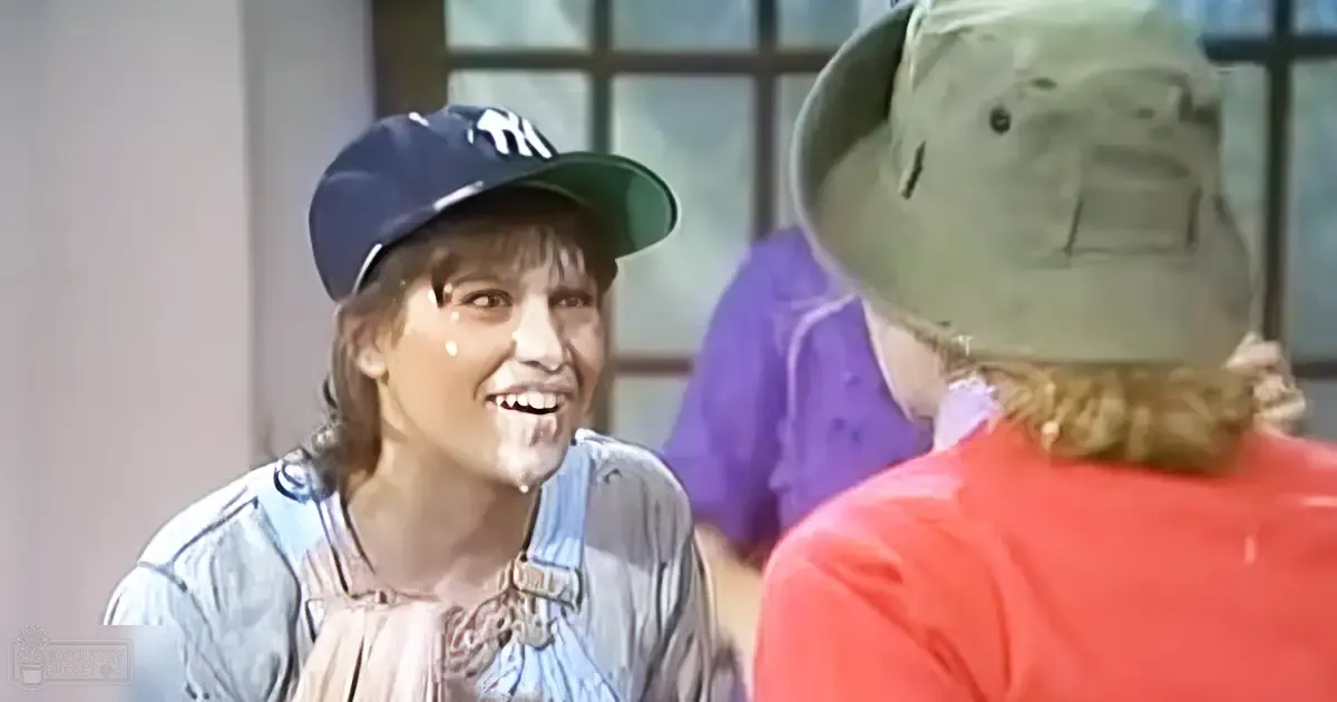 Young adult male wearing a dark blue baseball cap with 'TH' and light-colored shirt/jacket with patterned design, older adult in red shirt/beige bucket hat indoors near window panes.