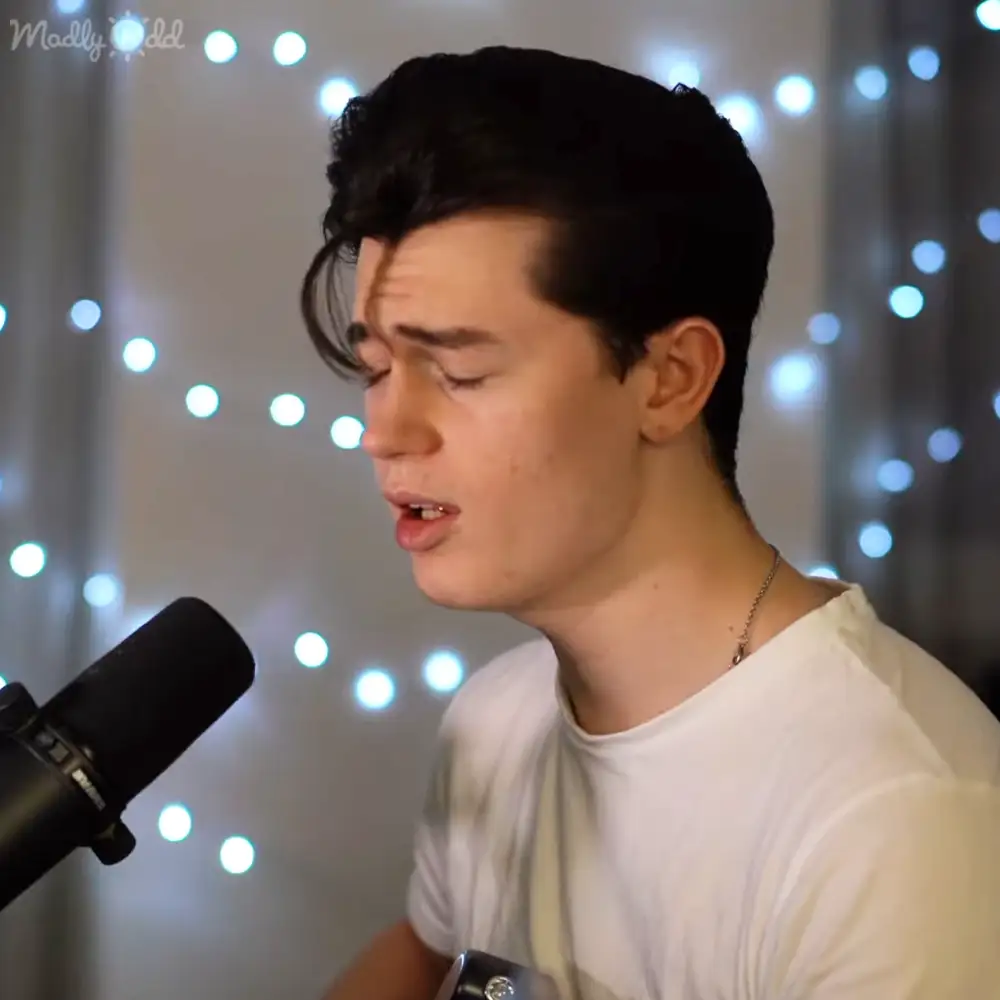 A young adult male wearing a white, relaxed fit crew neck t-shirt with short sleeves and a thin chain necklace performs in front of a microphone with string lights as background decor.