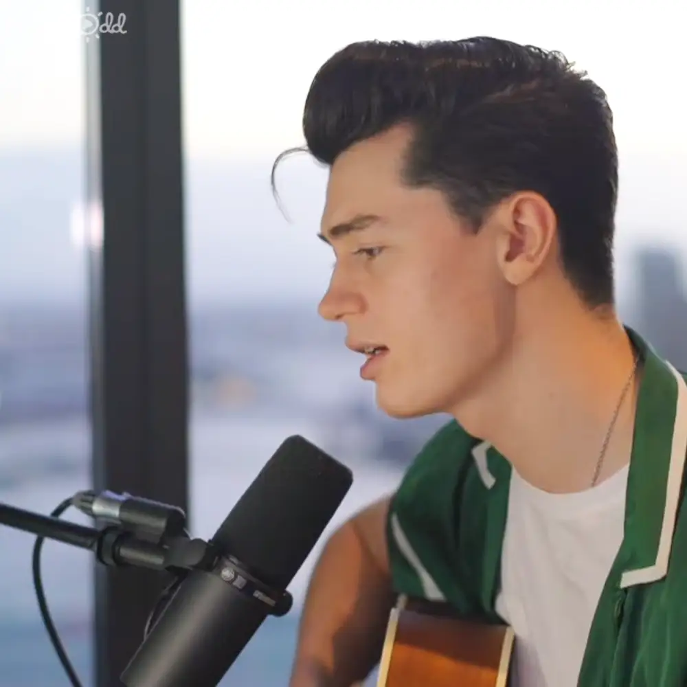 Young adult male in a green and white jacket singing into a microphone with a cityscape visible through large windows, wearing a necklace.