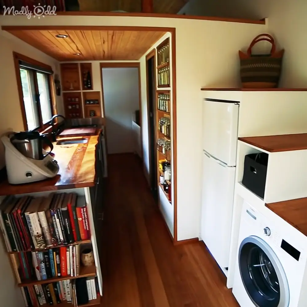 Compact kitchen area with wooden floor, white refrigerator, washing machine, shelves of books and bottles, countertop items like coffee maker and cutting board, natural light from window. Indoors in a home or apartment setting.