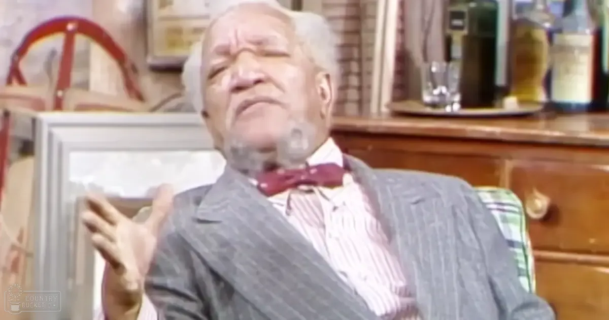 An older adult male in a pinstriped suit jacket over a white and pink striped dress shirt with a red bow tie, seated at a kitchen counter with a tray of glasses and bottles of alcohol, suggesting formal or semi-formal attire from the mid-20th century style.