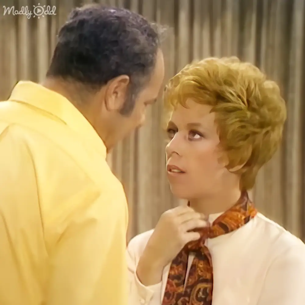 A woman in a patterned blouse stands beside an adult male singer wearing a bright yellow shirt against a backdrop suggesting a stage or studio environment.
