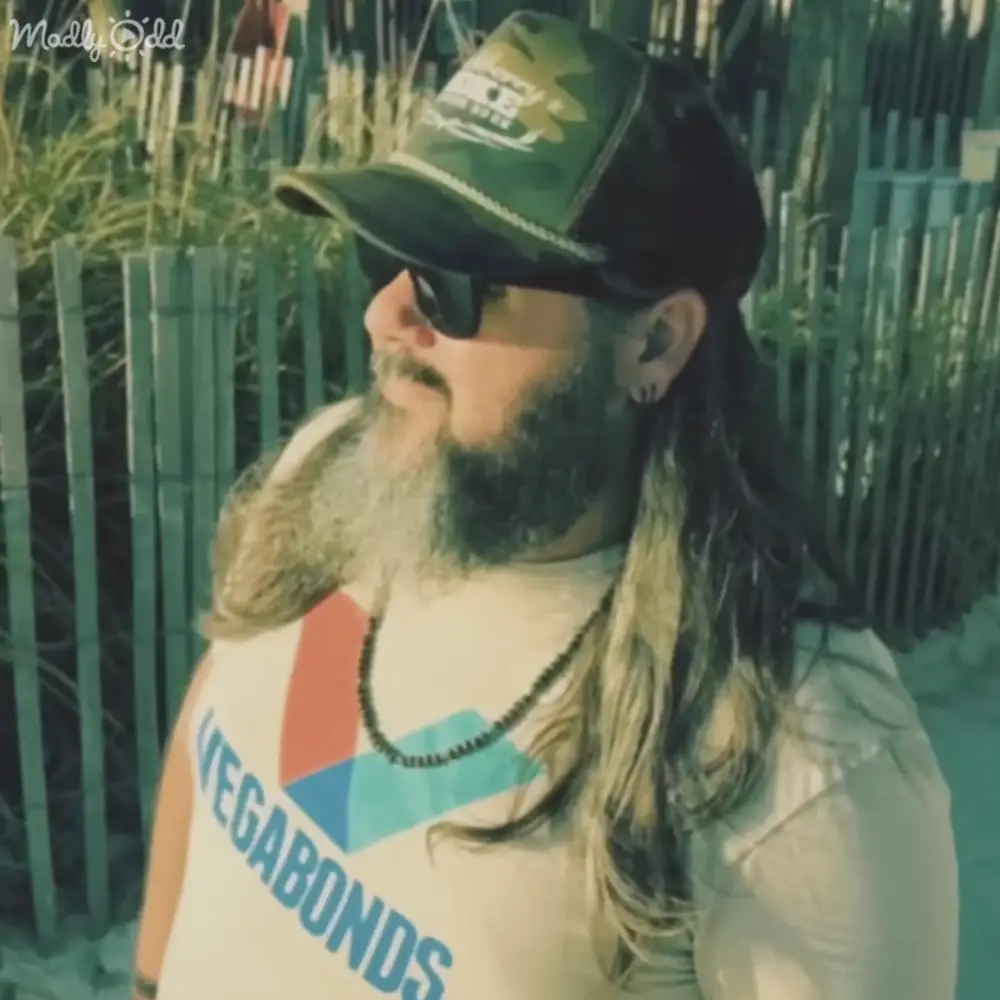 A middle-aged to older adult male wearing a white 'WEGABONDS' tank top with red and blue shapes, camouflage baseball cap, sunglasses, and a chain necklace outdoors near a green fence in bright daylight.
