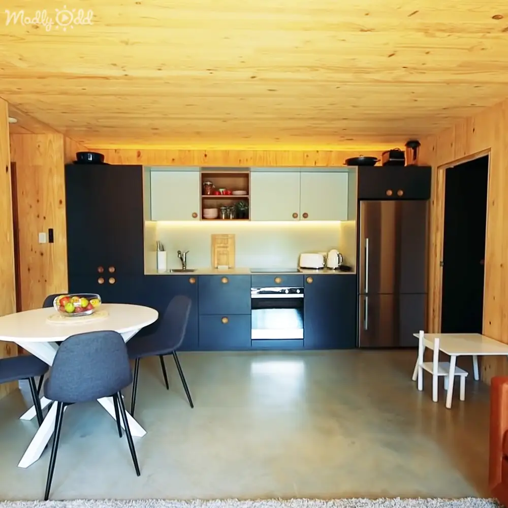 A modern kitchen space with a dining area with a white round table surrounded by dark blue chairs in an indoor setting with natural light streaming through.