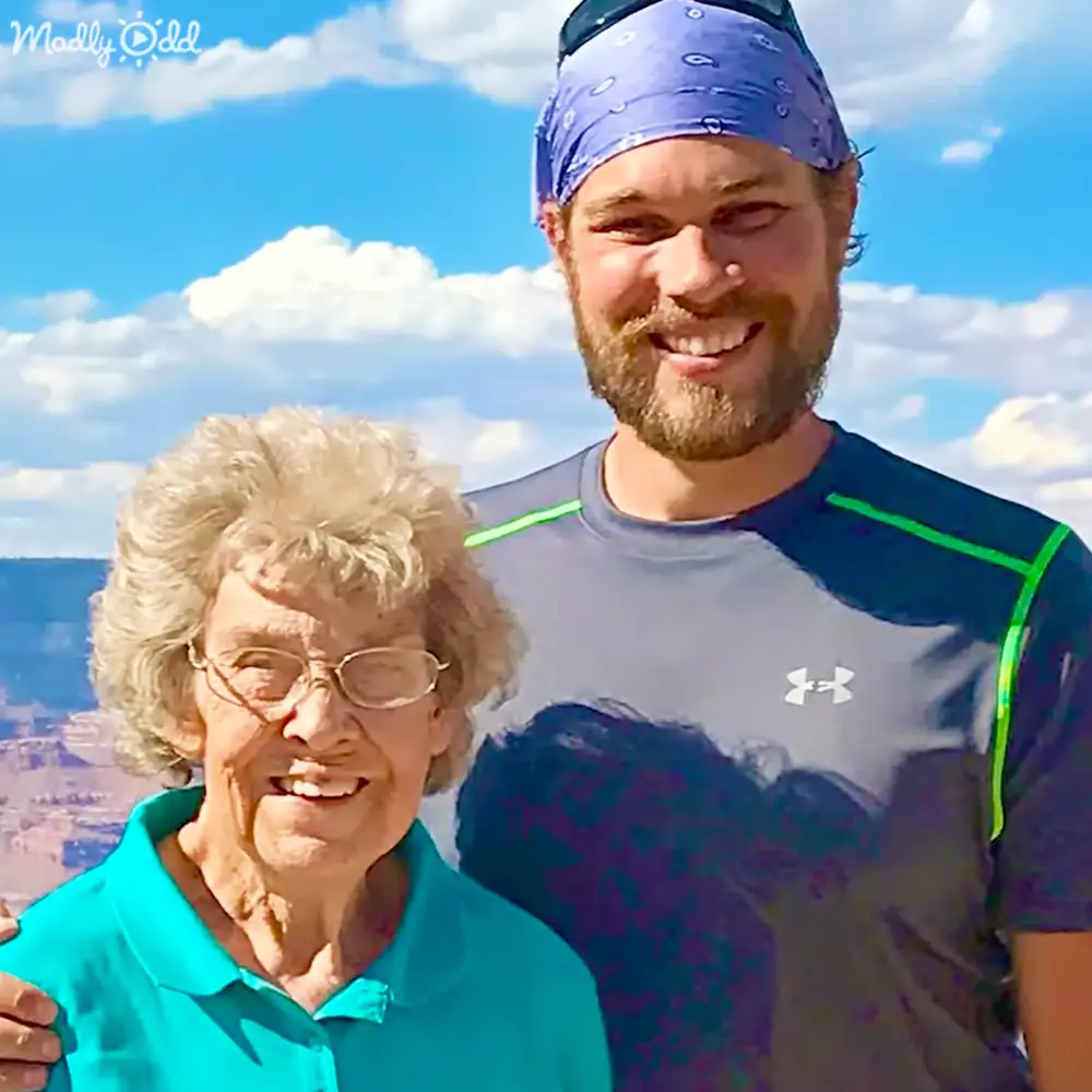 A young adult male and an older adult female outdoors against a scenic background with blue skies and clouds.