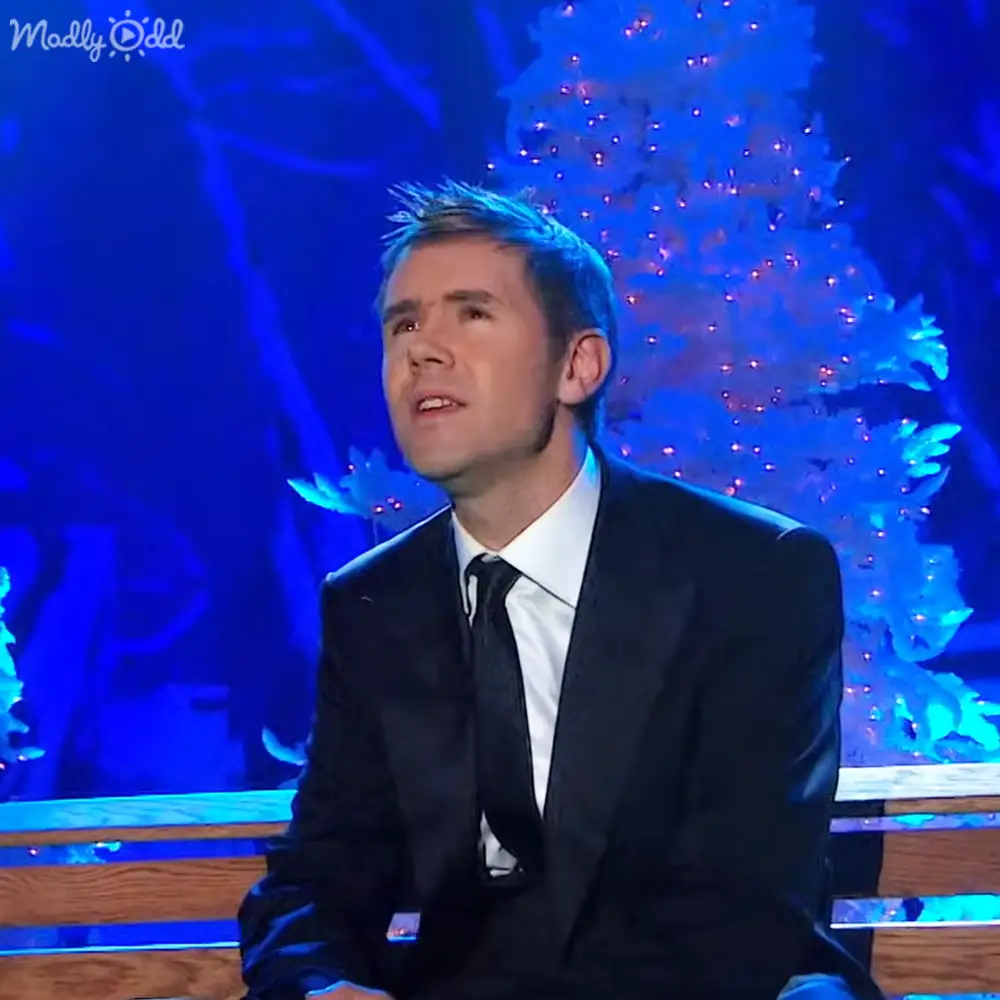 A young adult male wearing a dark tailored suit with white dress shirt and black tie, seated in front of a decorated Christmas tree in an indoor television studio setting.