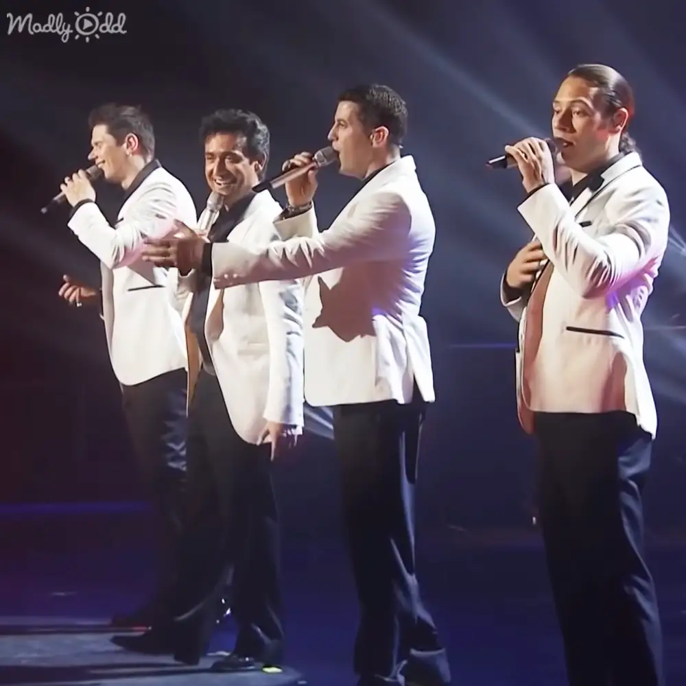 A group of adult male performers in white blazers and black pants on an indoor stage with dramatic spotlighting.