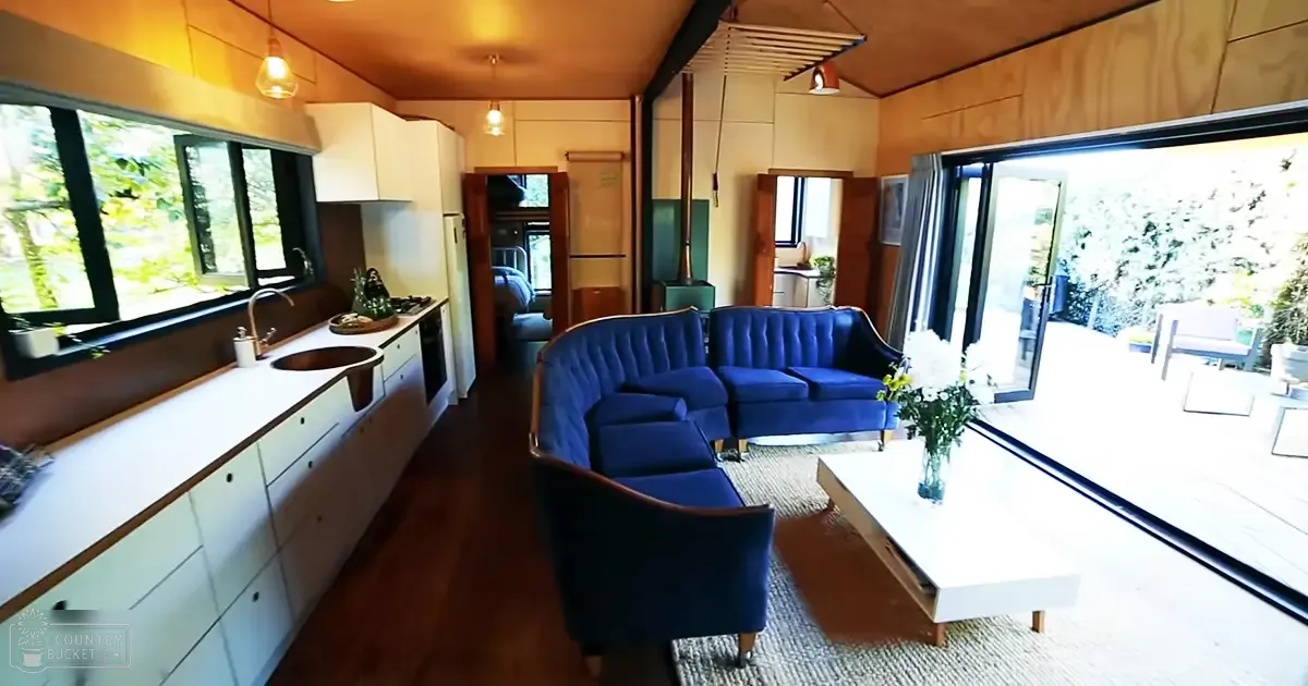 A modern home interior with a blue sofa, white coffee table with flowers, kitchen counter, and sliding glass door to an outdoor area with greenery.