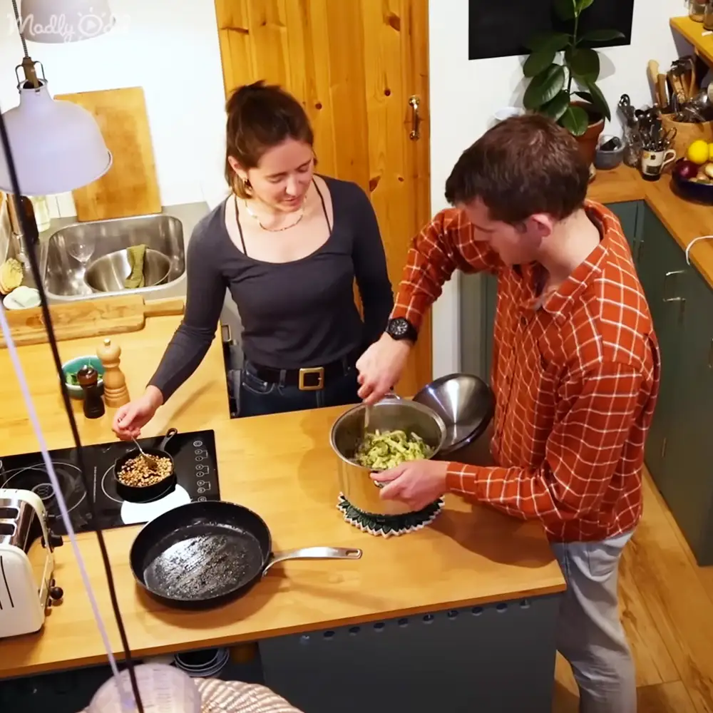 Two young adults preparing food in a kitchen with natural lighting, wooden furniture, and various kitchen utensils around. Season and time of day are ambiguous.