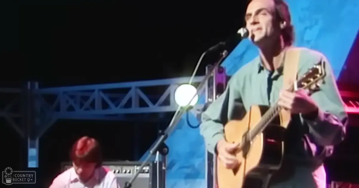 Middle-aged male musician performing live at an outdoor concert, wearing a light green long-sleeve shirt and dark pants, with white spotlights illuminating the stage in the background.