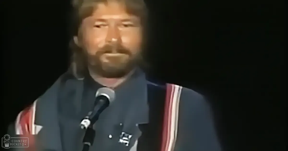 A middle-aged man in a 1970s-style jacket performs on stage at an indoor concert or event.