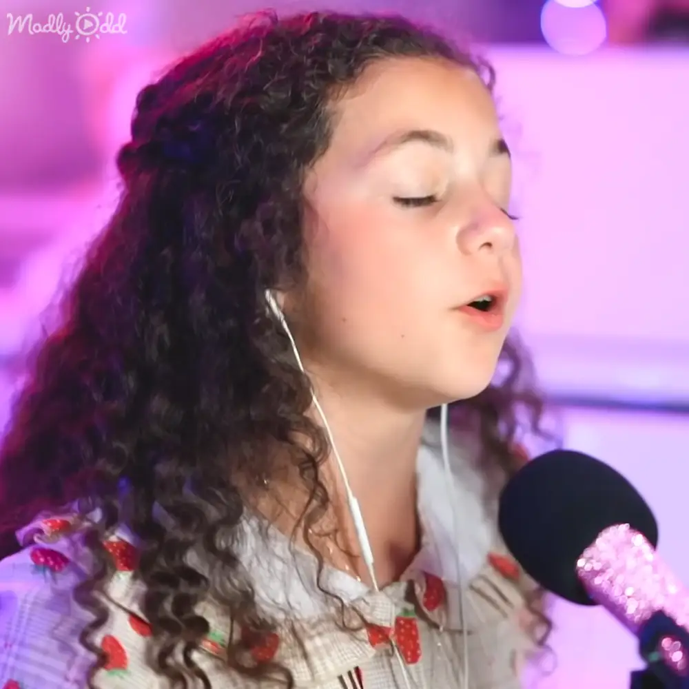 A young child wearing a white blouse with strawberry patterns sings into a microphone on stage under purple lighting.