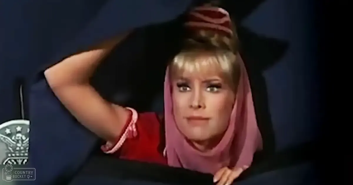 A woman with a red high-necked garment and pink headscarf stands against a dark, dimly lit backdrop.
