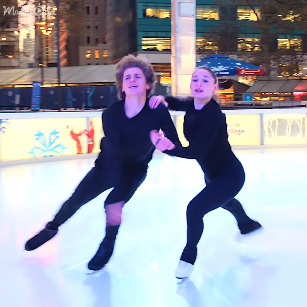 A young couple skating at a night-time urban ice rink, surrounded by illuminated buildings with signs.